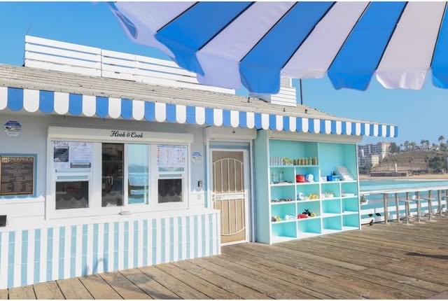 Hook & Cook’s white pier shack with blue striped awnings and an aqua display beside the Pacific Ocean.